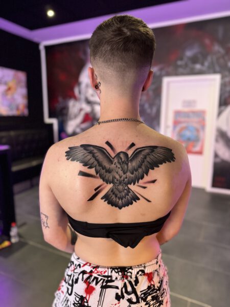 Rückenansicht einer Person mit einem großen, detaillierten Tattoo eines fliegenden Vogels auf der Haut. Die Person trägt ein schwarzes Oberteil und hat eine kurze Frisur. Der Hintergrund zeigt eine künstlerische Umgebung mit bunten Elementen.