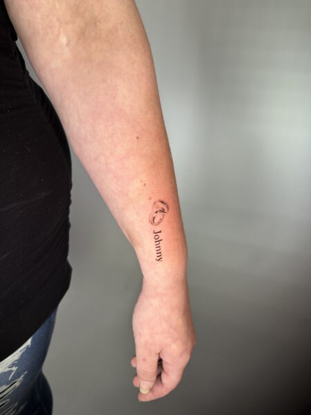 Tattoostil Fineline - Schriftzug "Jonny" und Eheringe auf einem Unterarm.